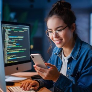 happy-young-woman-looking-her-smartphone-texting-while-coding-her-computer (1)